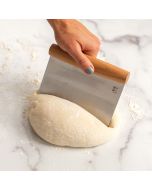 A person uses a dough scraper to cut a dough ball on a wooden countertop.