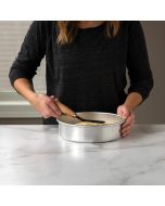 A woman holds a spatula over a metal pan, preparing food in a kitchen setting.