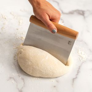 A person uses a dough scraper to cut a dough ball on a wooden countertop.