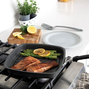 A searing grill pan on a stove holds salmon and asparagus, garnished with a slice of lemon.