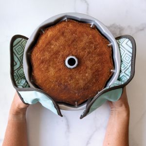 A person holding a freshly baked cake in a round pan, smiling and presenting it to the camera.