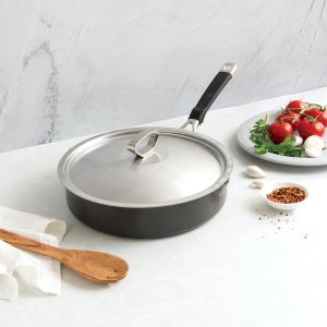 A pan with a wooden spoon beside a bowl filled with fresh strawberries on a kitchen countertop.