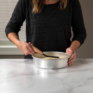 A woman holds a spatula over a metal pan, preparing food in a kitchen setting.